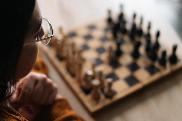 Closeup rear view of thoughtful young woman in elegant eyeglasses thinking about chess move sitting in dark room selective focus