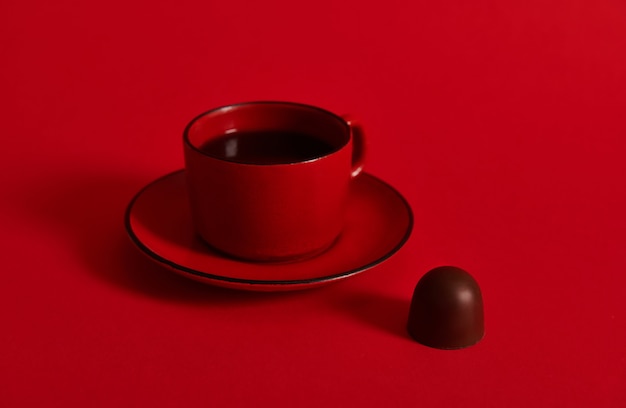 Closeup of a red ceramic cup of coffee on a saucer and a chocolate praline on red surface with copy space . Concept of chocolate day