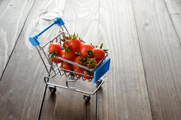 Closeup red shiny cherry tomatoes in the blue shopping cart on dark wooden background