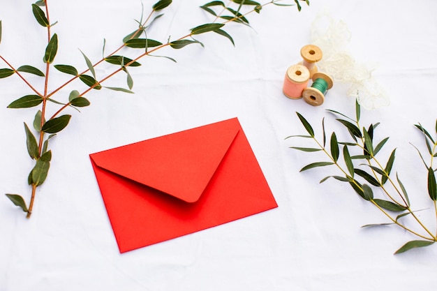 Closeup rode envelop omringd door verse eucalyptus takken en verschillende kleine draadspoelen Van bovenaf foto met kopie ruimte op witte achtergrond Verzenden groeten concept