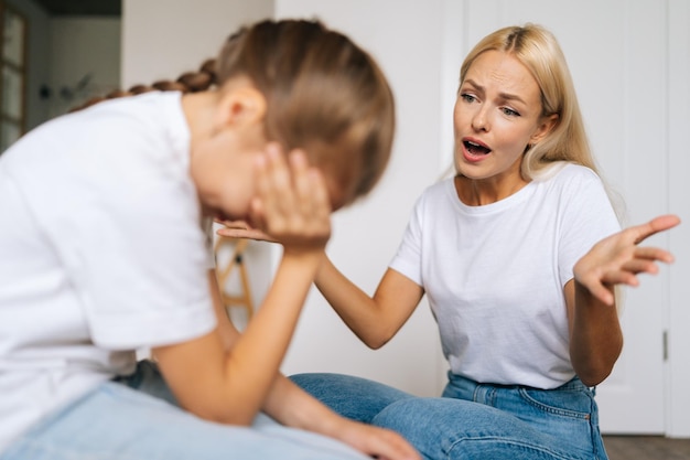 Closeup of sad depressed little girl crying covering face with palm while angry young mother scolding scream at stubborn difficult little child daughter