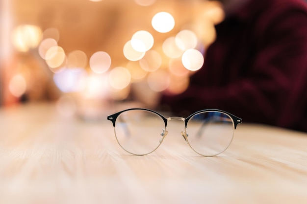 Closeup selective focus of fashion glasses lying on wooden\
table on blurred bokeh background closeup of black eyeglasses on\
desk with bokeh christmas light