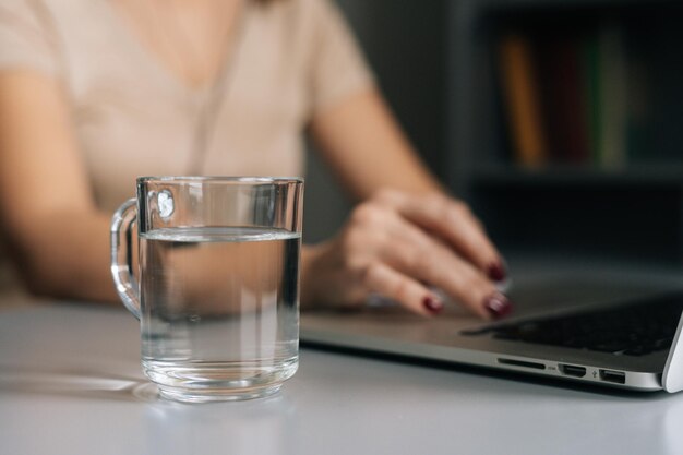 Closeup selective focus of glass water on table where unrecognizable business woman working on laptop computer at home office