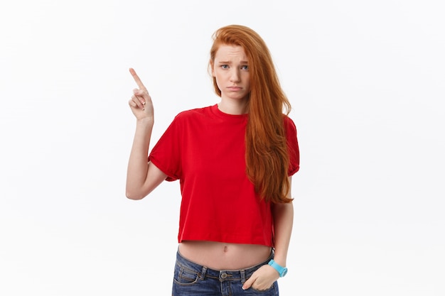 Closeup of serious strict young woman wears red shirt looks stressed and pointing up with finger isolated over white
