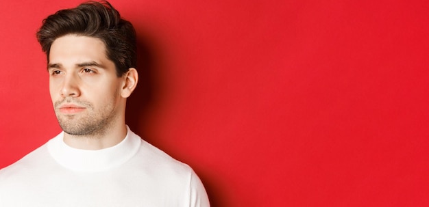 Closeup of serious and thoughtful man in white sweater looking left standing against red background