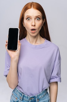 Primo piano di una giovane donna scioccata in abiti casual che tiene in mano un telefono cellulare con schermo mobile vuoto nero