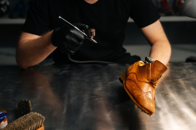 Photo closeup of shoemaker in black gloves spraying paint of light brown leather shoes