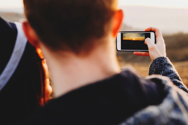 スマートフォンを使用して田舎の素晴らしい夕日の写真を撮る匿名のカップルのクローズアップショット。日没の写真を撮る作物のカップル