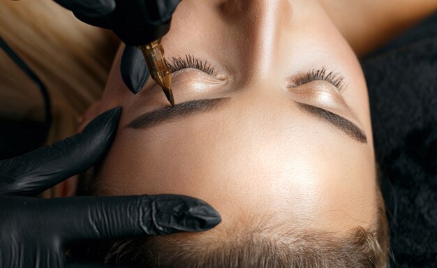 Closeup shot of a beautician in black gloves applying permanent brow makeup to a blonde woman. Space for text