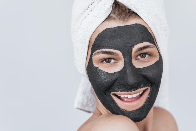 Closeup shot of beautiful smiling young female doing beauty treatment on her face clean skin with black organic clay looks at the camera with toothy healthy smile Woman applying organic facial mask