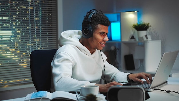 Closeup shot of black gamer in white hoodie and with headphones playing games on computer in the evening