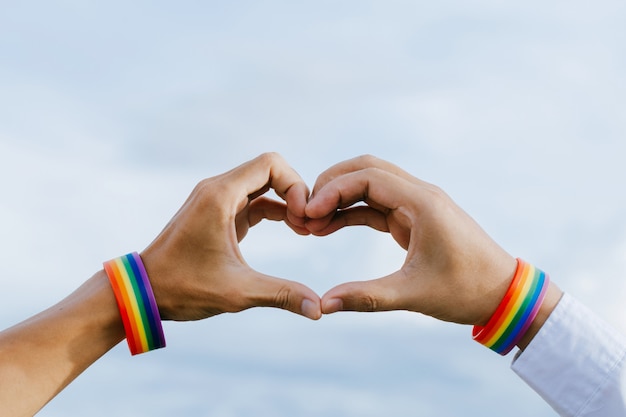 Primo piano di una coppia gay che si tiene per mano con un braccialetto arcobaleno fatto mani formando una forma di cuore