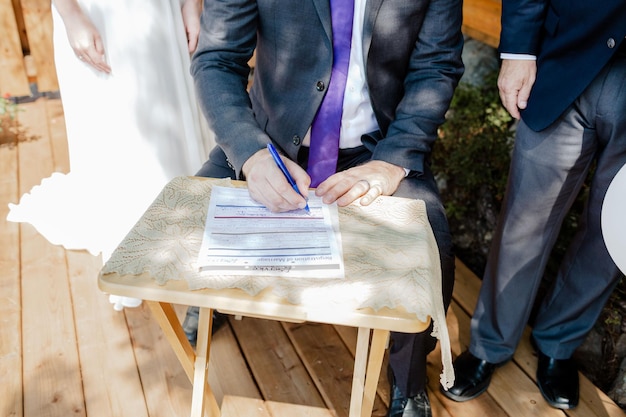 Foto scatto ravvicinato di uno sposo che firma un contratto di matrimonio su un tavolo di legno in una giornata di sole