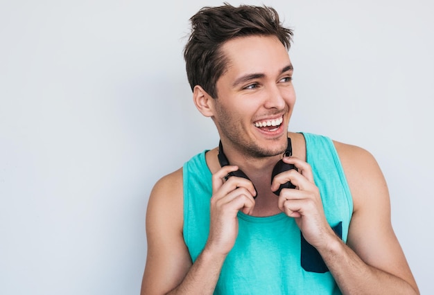 Colpo del primo piano dell'uomo caucasico alla moda bello che indossa la canotta colorata dei pantaloni a vita bassa che sorride mostrando i denti bianchi che posano contro il fondo bianco della parete con le cuffie sul suo collo che distoglie lo sguardo