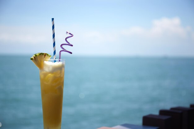 Closeup shot of an icy pineapple juice with swizzle sticks on a blurred sea background and blue sky