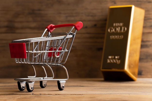 Closeup shot of a large shiny gold bar near the tiny shopping cart with wooden wall background