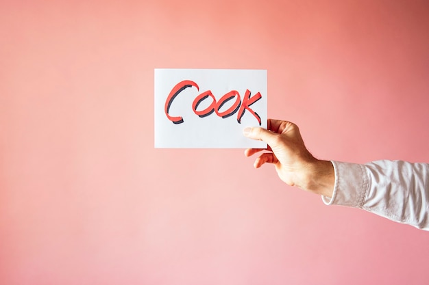 A closeup shot of a person holding a paper with the word "COOK" on a pink wall