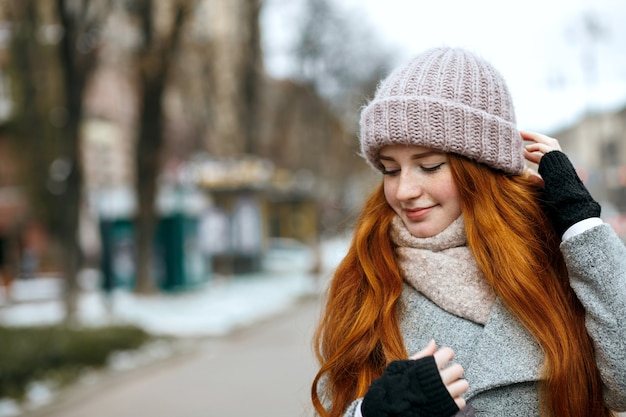 Closeup shot of pretty ginger girl with long hair wearing knitted cap and scarf walking at the city. Space for text