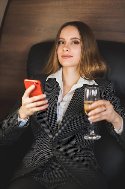 Closeup shot of pretty self assured caucasian young businesswoman in stylish suit, with makeup, long hair sitting in private jet with glass of champagne and mobile phone in hands.