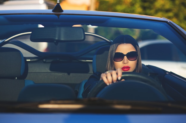 Colpo del primo piano della donna castana alla moda con le labbra rosse che indossano occhiali da sole che guidano un cabriolet. spazio per il testo