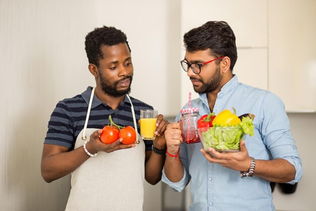 밝은 부엌에 서서 신선한 유기농 야채와 과일 음료가 든 안경을 들고 있는 두 명의 다민족 친구의 근접 촬영. 건강 식품, 채식주의, 요리 개념.