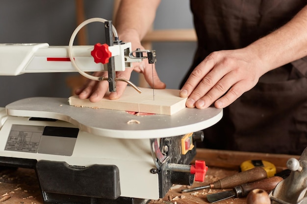 Closeup shot of unknown unrecognizable faceless man carpenter wearing brown apron working in his joinery works with an electric jigsaw making wood products