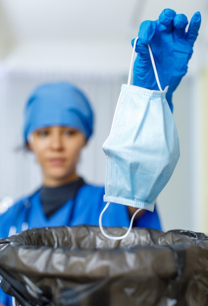 Closeup shot of used face mask was dumped into garbage bag trash can by female happy smiling doctor in blue hospital uniform rubber gloves and stethoscope in blurred background after pandemic ended.