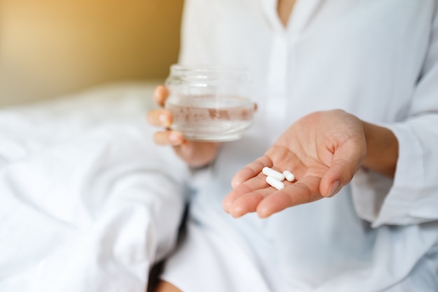 Closeup  of a sick woman holding white pills and a glass of water while sitting on a bed