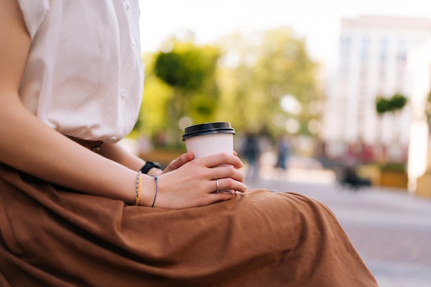 Фото Вид сбоку крупным планом обрезанный снимок неузнаваемой молодой женщины, держащей в руках бумажную чашку с кофе на вынос, сидящей на скамейке на городской улице