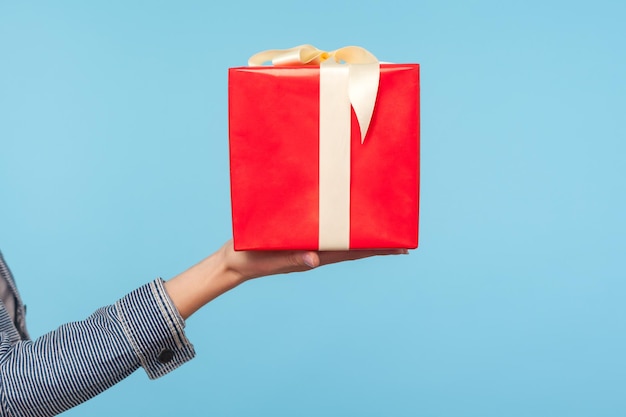 Closeup side view of female hand in denim clothes holding big red gift box, woman arm showing wrapped present, bonuses and congratulations on birthday. indoor studio shot isolated on blue background