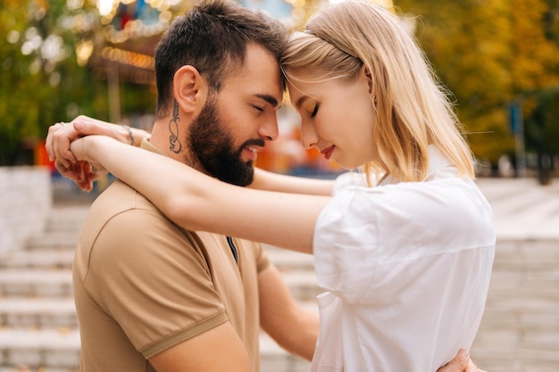 Closeup side view of happy young couple in love touching each other foreheads hugging standing in amusement park in summer day