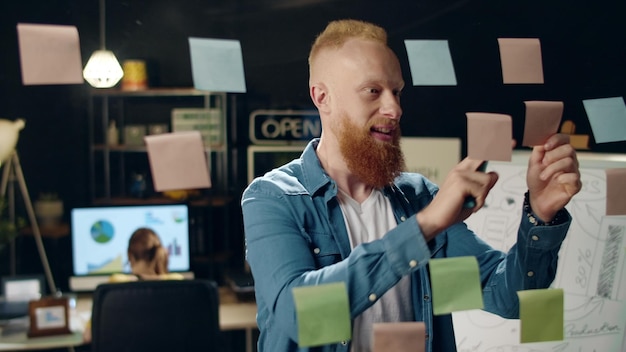 Closeup smiling hipster guy sticking notes on glass board on night office