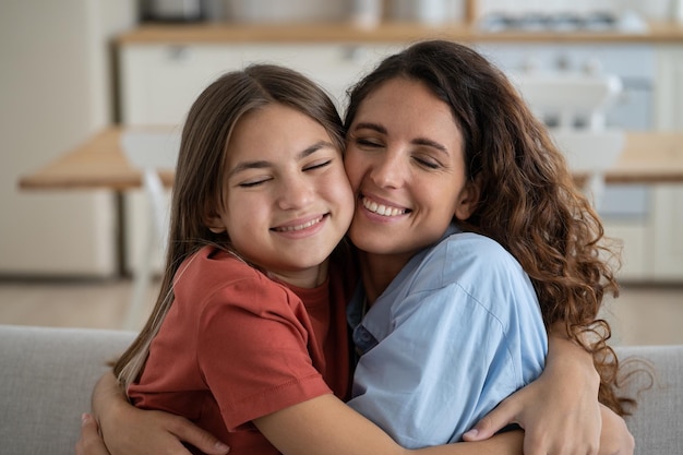 Closeup smiling positive mother and daughter hugging each other tightly sits on sofa in apartment