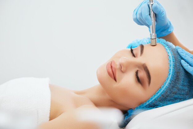 Closeup of smiling woman in blue cap lying on couch in cosmetological clinic with closed eyes. Doctor's hands in blue gloves doing procedure of microdermabrasia on her face