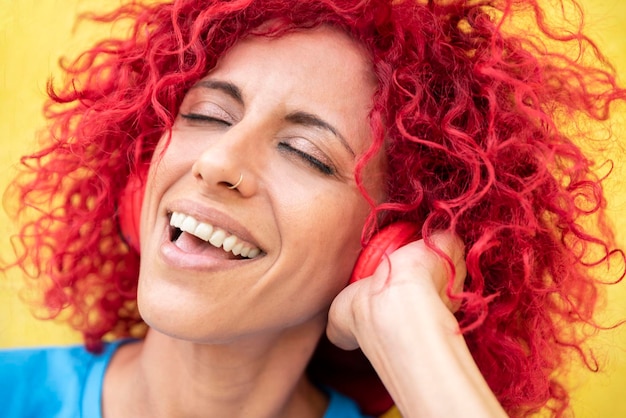 Closeup of a smiling young latin woman with red afro hair wearing a blue tshirt listening to music with her eyes closed and singing holding headphones with one hand on a yellow background