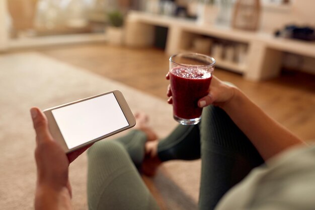 Closeup of sportswoman using smart phone and having a glass of fruit smoothie at home