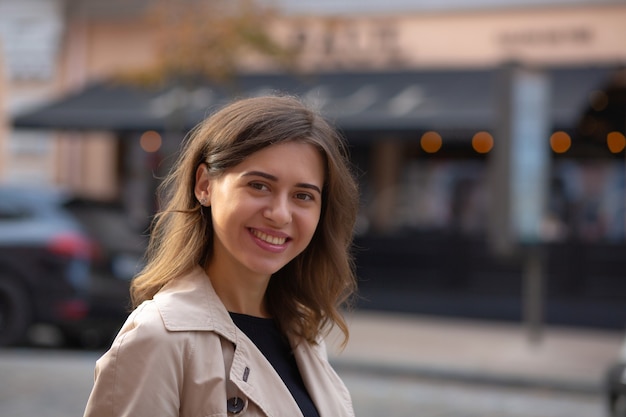 Closeup street shot: cheerful elegant woman with nude makeup wears beige spring coat. Space for text