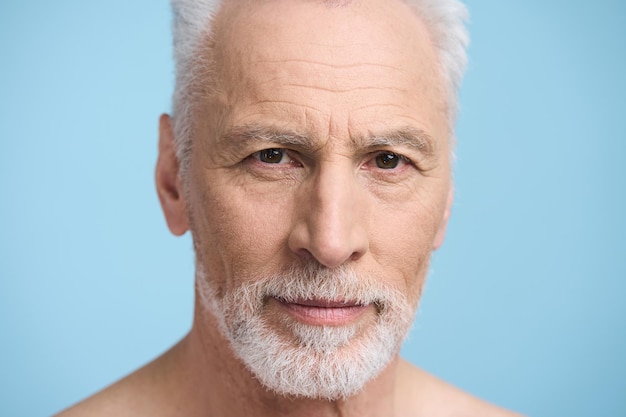closeup studio portrait face of caucasian handsome gray haired bearded senior man 60s looking at camera isolated on blue background healthy lifestyle male beauty barbershop skin and body care