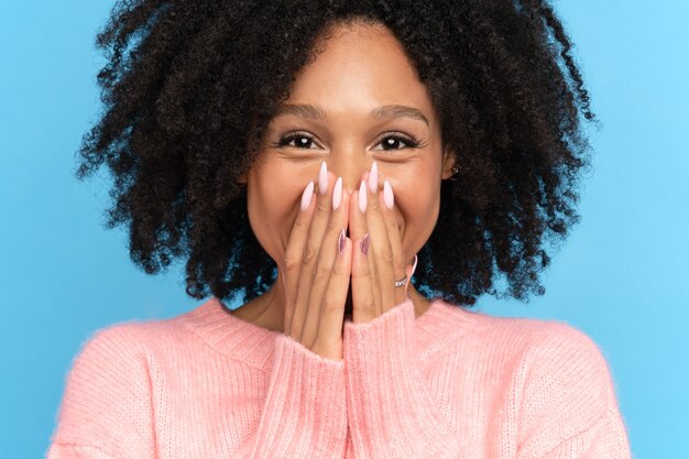 Closeup studio portrait of happy African girl. Laughing dark skinned millennial woman looking at camera. Cute positive mixed race female smiling covering her mouth and lips with hands, blue background