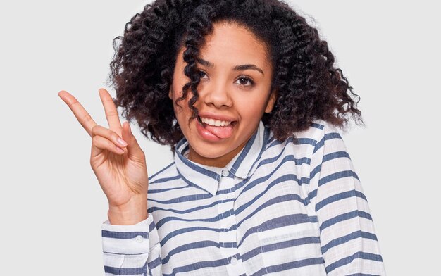 Closeup studio portret van positieve Afro-Amerikaanse jonge vrouw die lacht in grote lijnen vredesgebaar tonen en tong toont terwijl ze naar de camera kijkt die tegen een witte studioachtergrond staat