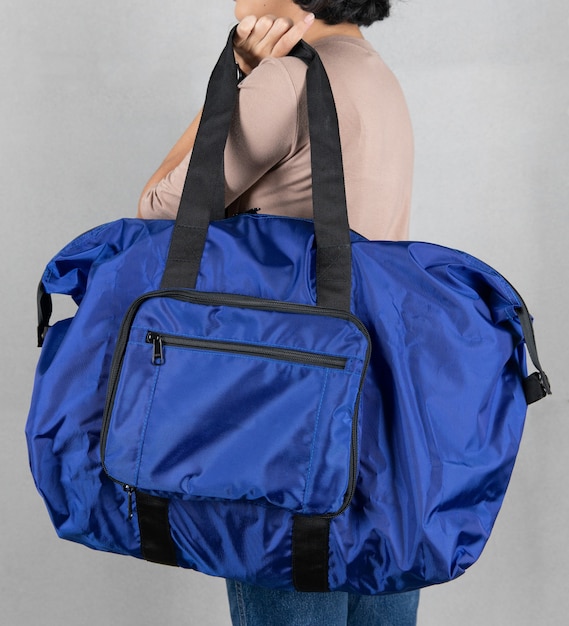 Closeup studio shot of female model in brown long sleeve shirt\
and jeans stand holding handle of fashionable trendy urban big gray\
unisex handbag with long shoulder strap in front of grey\
background.