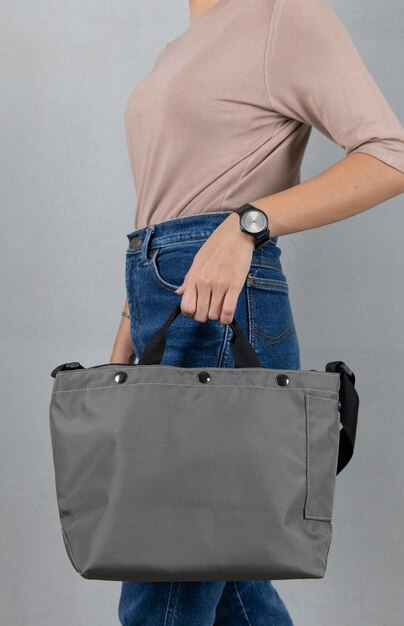 Closeup studio shot of female model in brown long sleeve shirt and jeans stand holding handle of fashionable trendy urban big gray unisex handbag with long shoulder strap in front of grey background.