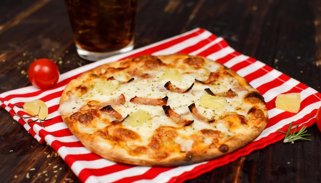 Closeup studio shot hot oven baked tasty delicious Italian homemade ham pineapple traditional Hawaiian bread crust round pizza decorated on red white stripe cloth with tomato and herbs on wood table