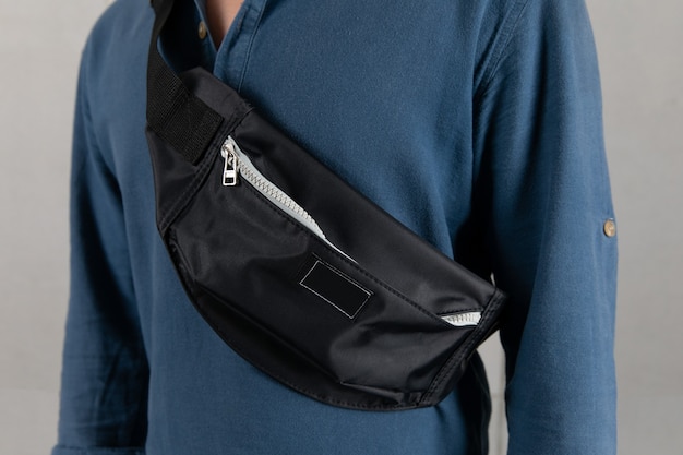 Closeup studio shot of male model in blue long sleeve shirt\
hanging trendy urban small black crossbody strap casual fanny pack\
bag in front gray background.