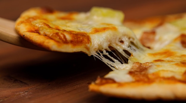Photo closeup studio shot using wooden scoop peel picking up hot oven baked tasty delicious italian homemade ham and pineapple hawaiian soft bread crust pizza piece from wood plate pan on restaurant table