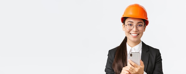 Closeup of successful female chief engineer construction architect in safety helmet and business suit glasses looking at camera satisfied smiling pleased as using mobile phone