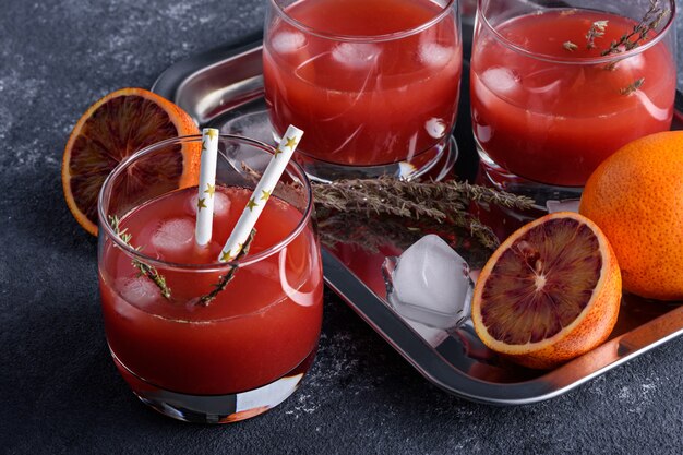 Closeup summer citrus juice from red oranges with ice in glasses on tray on gray background