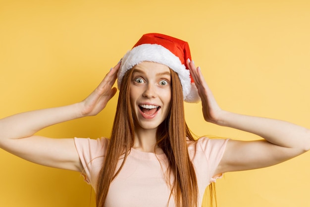 Closeup of surprised happy young caucasian redhead freckled female wearing Santa hat keeping hands on head, amazed by big Christmas discounts in the store, standing on yellow background.