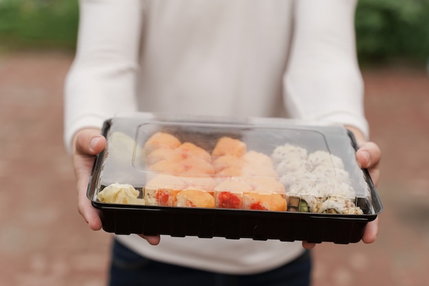 Closeup sushi in box healthy food delivery online service. Man in white clothes gives sushi set to you. Japanese cuisine rolls, soy sauce, wasabi.