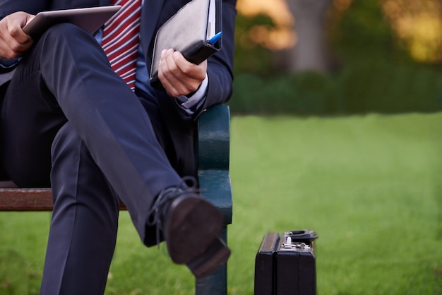 Photo closeup tablet and business man on bench park and planning with lunch break and professional employee outdoor and consultant with suitcase and technology for ideas and shoes with fashion or relax